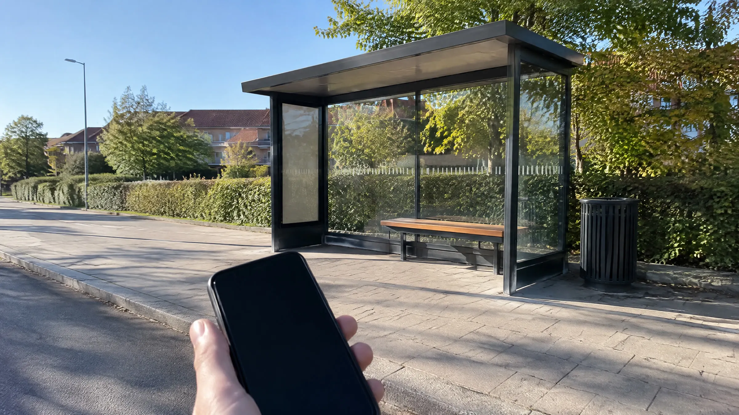 En hånd holder en mobiltelefon med sort skærm foran et tomt busskur ved en vej med høje hække