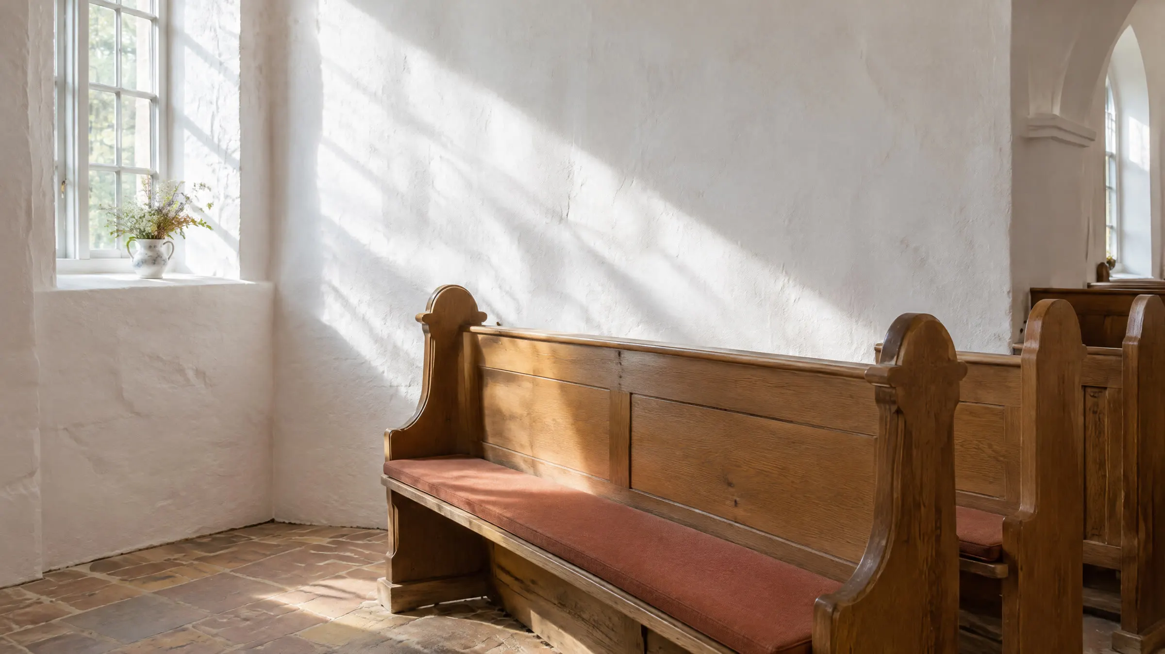 En kirkebænk af træ i en hvidkalket dansk landsbykirke med sollys på den bare væg