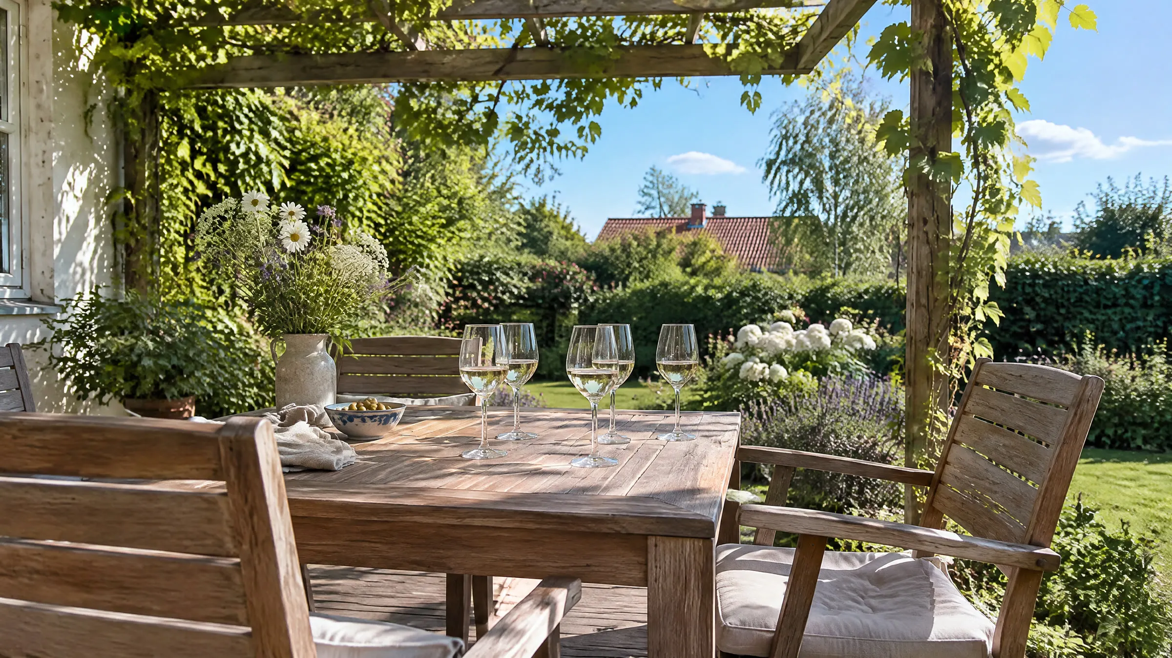 Fem glas hvidvin på et havebord i træ under en overgroet pergola i en solrig sommerhave